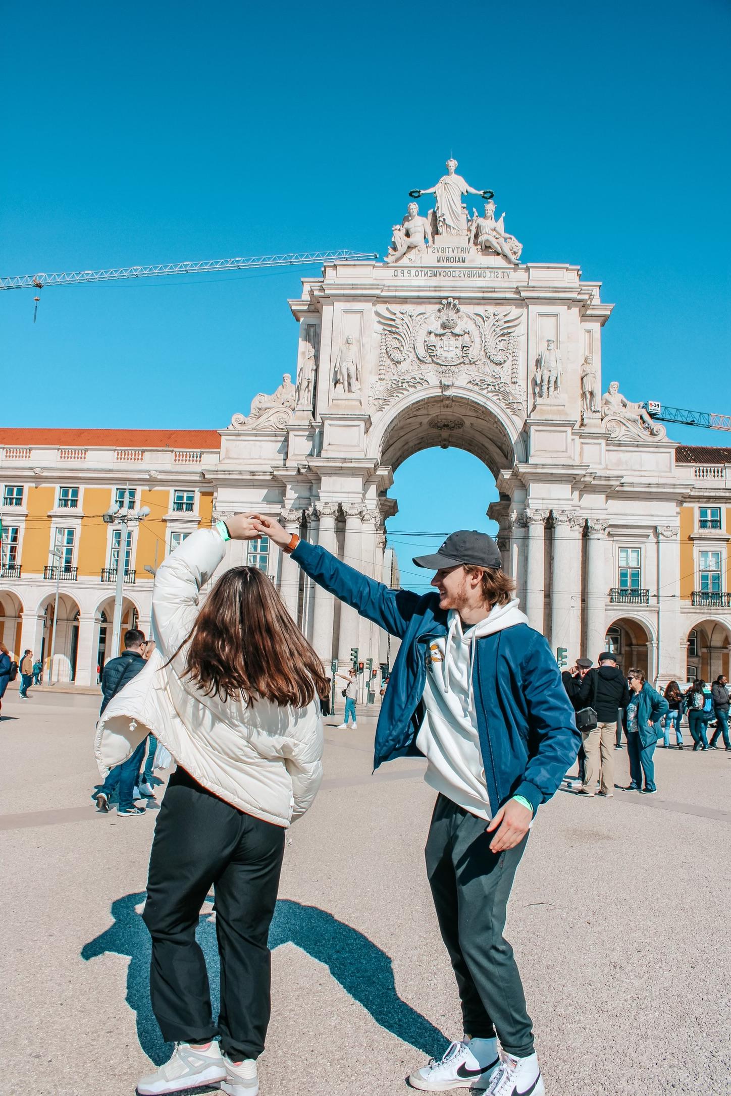 Woman in white jacket twirling with man in black baseball cap and blue jacket in front of Arco da Rua Agusta